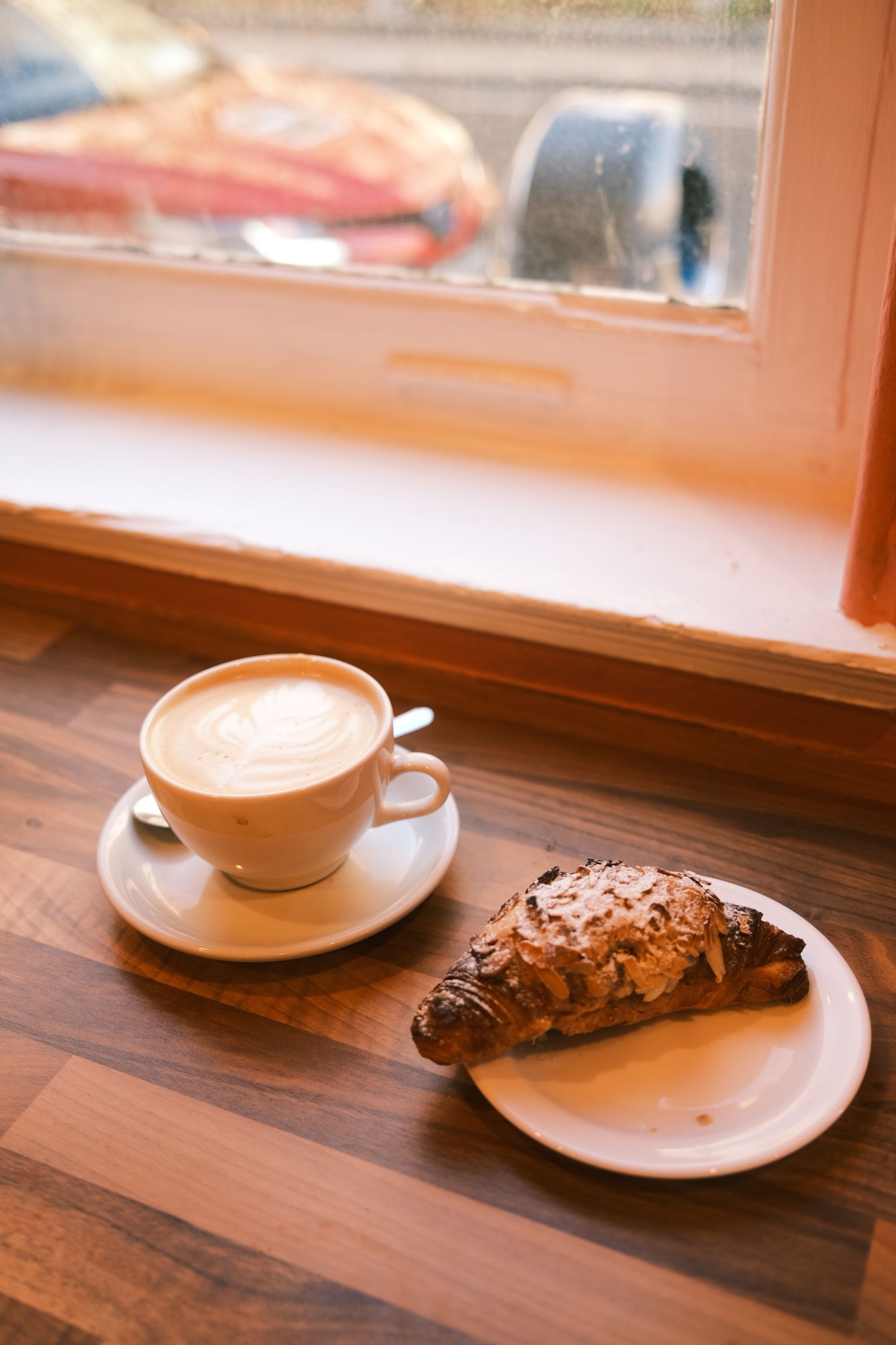 Cappuccino and golden almond croissant on a warm wooden table by a sunlit window