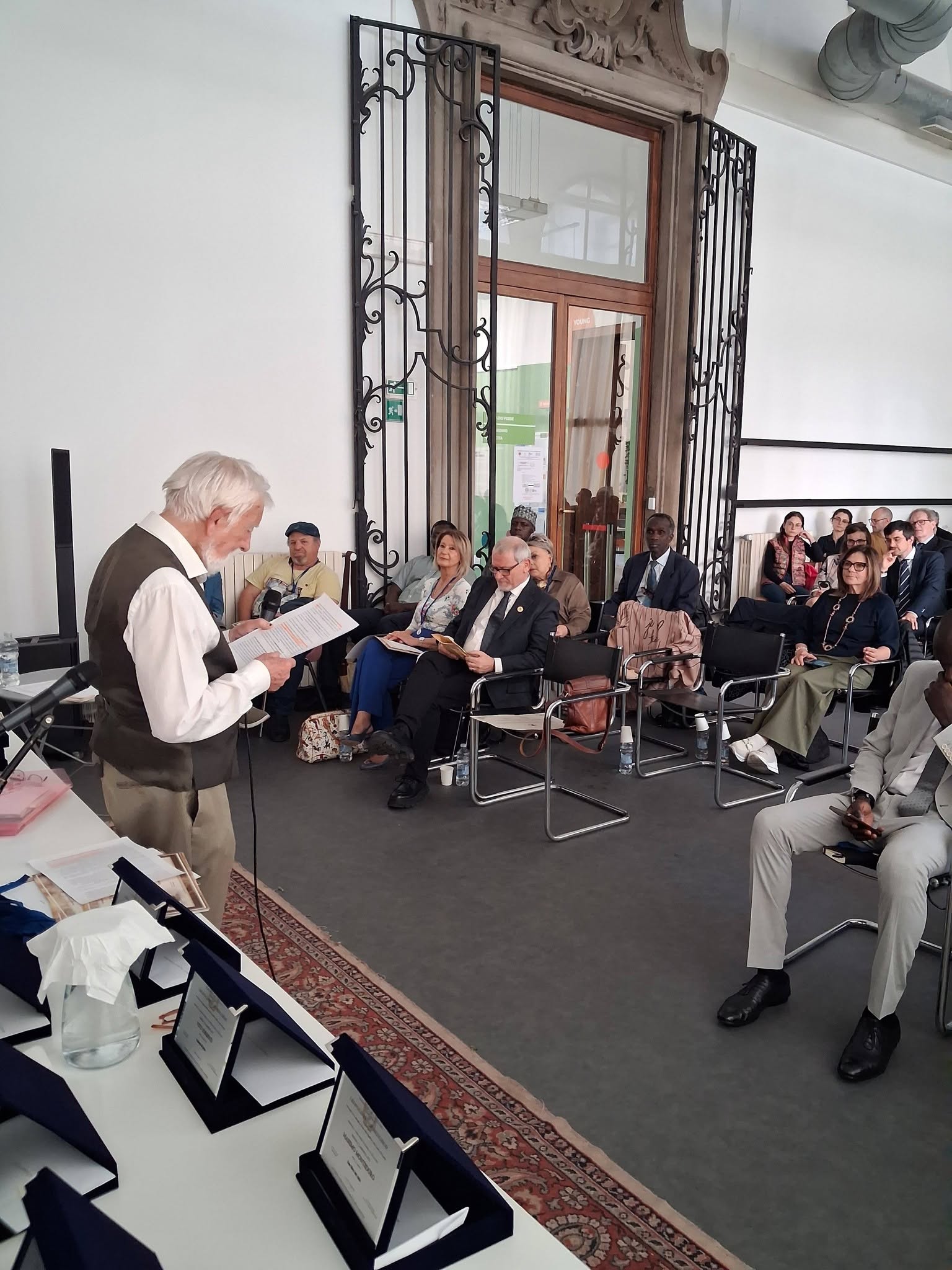 Un homme lit un texte devant le public assis dans la salle.