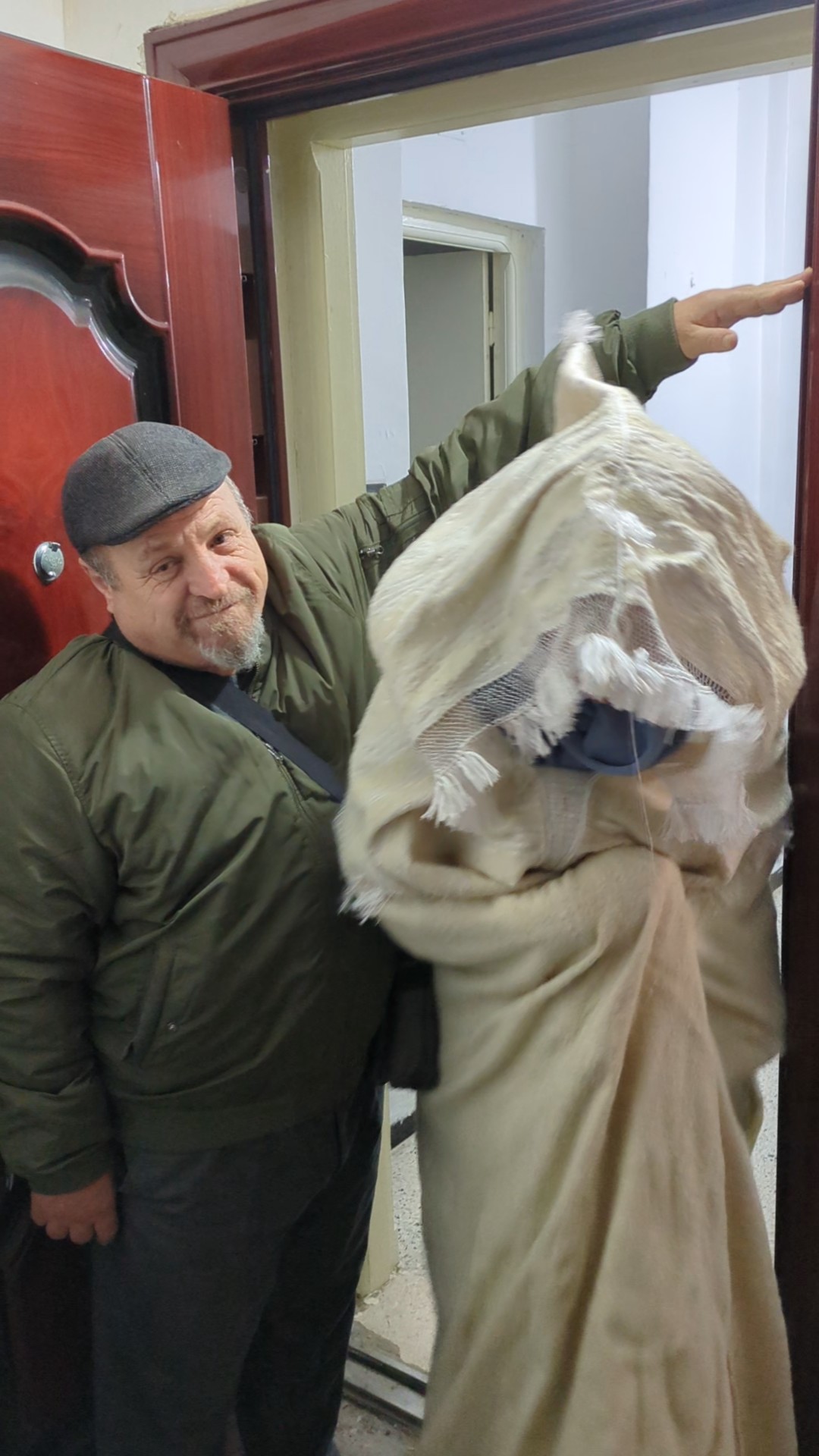 Un homme en casquette et veste verte auprès d’une personne portant un burnous kabyle blanc à capuche et à franges, dans l’encadrement d’une porte.